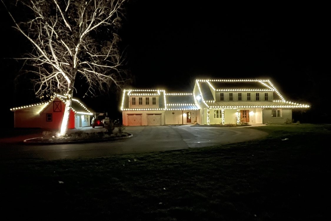 Colonial home with a red barn, roofline and dormers outlined in warm white lights, a bare tree wrapped to the branches