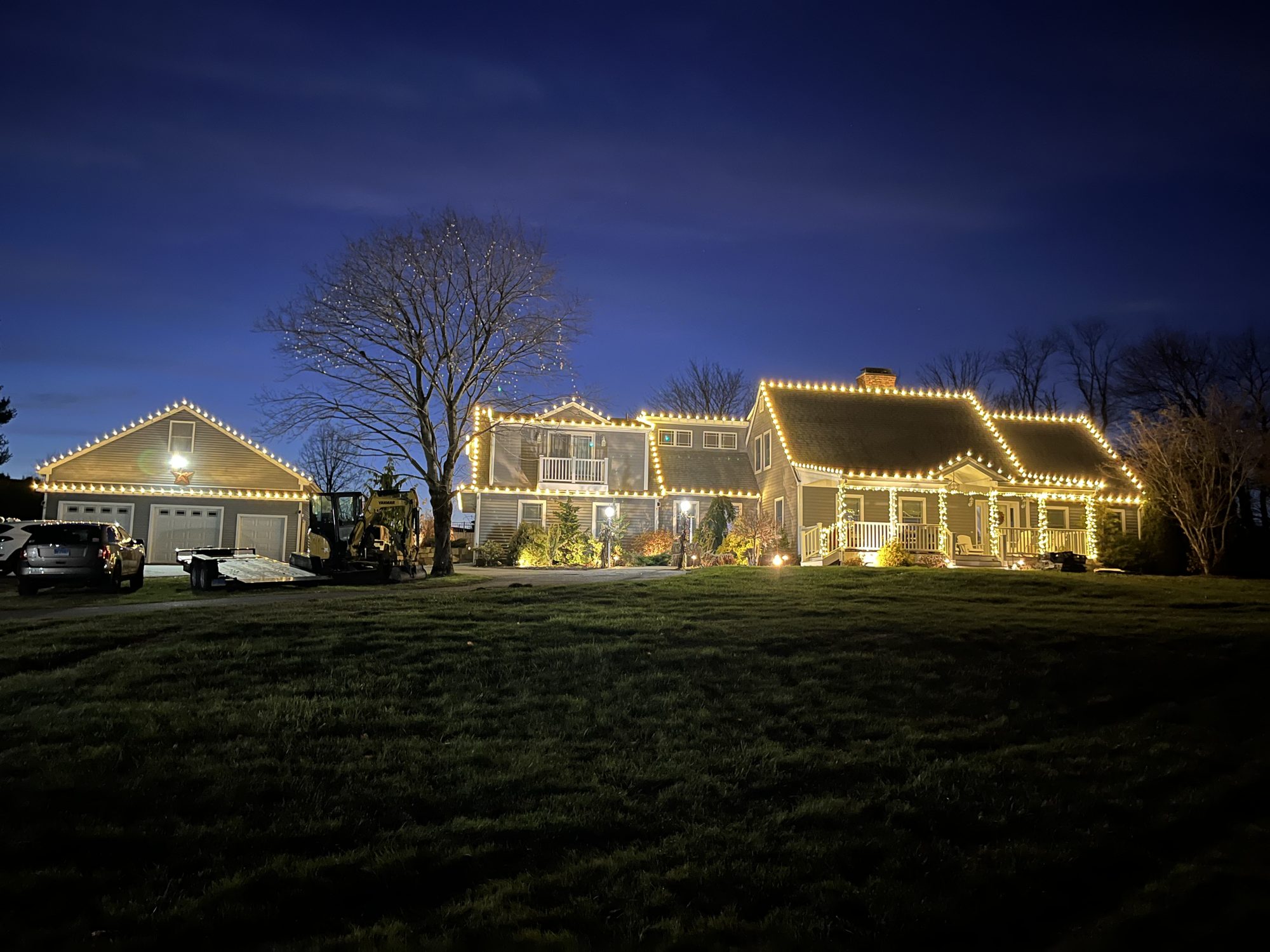 Home and detached garage outlined in warm white roofline lights at dusk, with a front tree and porch columns lit